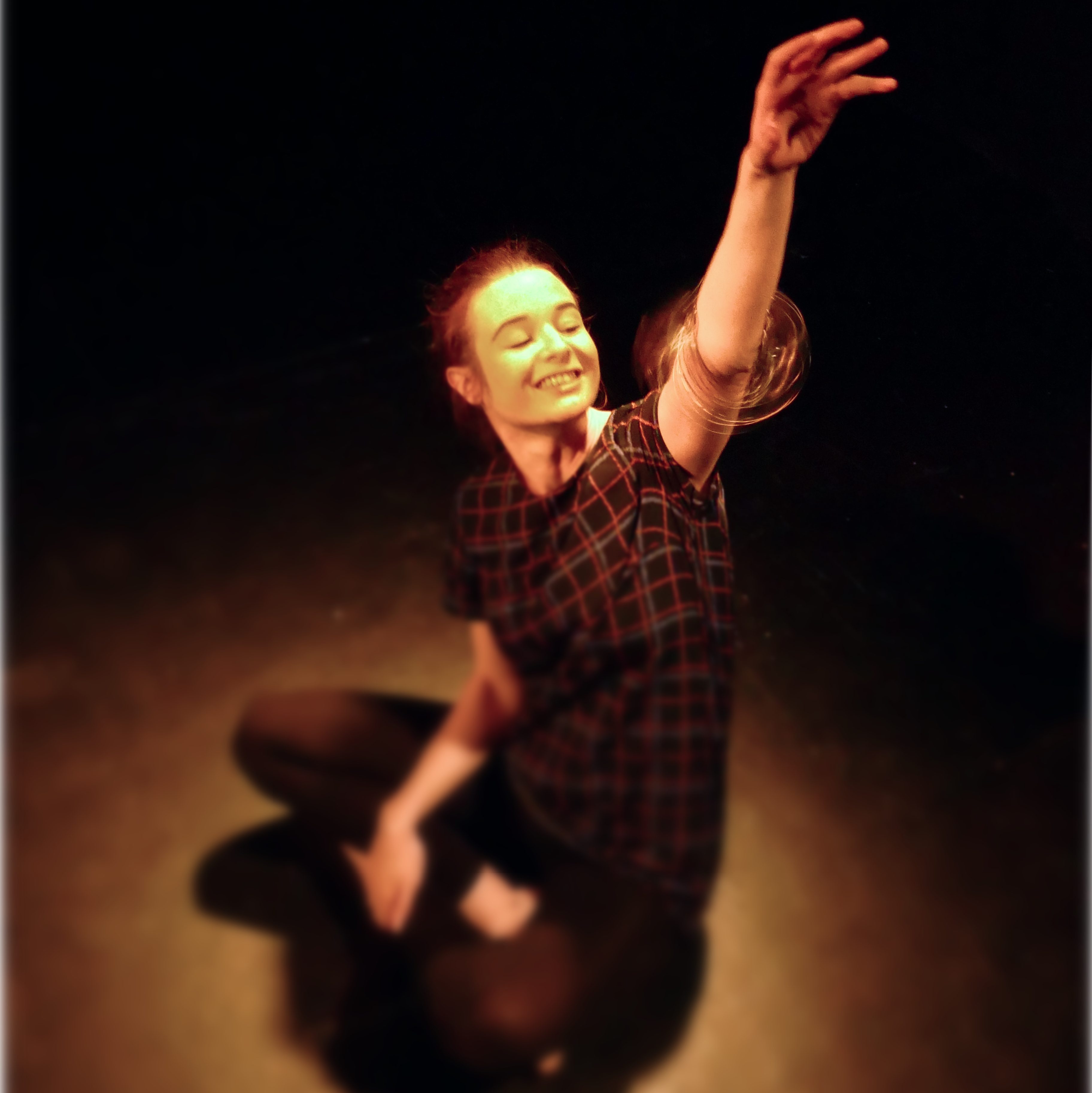 A white woman with brown hair, sitting in a spotlight with her arm in the air. There is a 'magic ring' stim device spinning around her arm and she is watching it and smiling