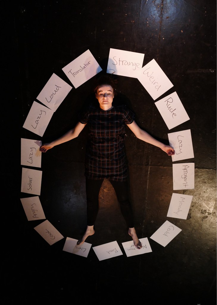A young white woman with brown hair tied back, lying on the floor, staring up at the ceiling, surrounded by pieces of paper that say negative words like rude, lazy, strange, weird, loud