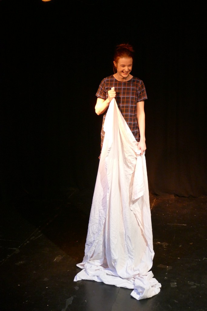 A young white woman with brown hair tied back, laughing whilst standing on a chair and holding up a sheet on a stage
