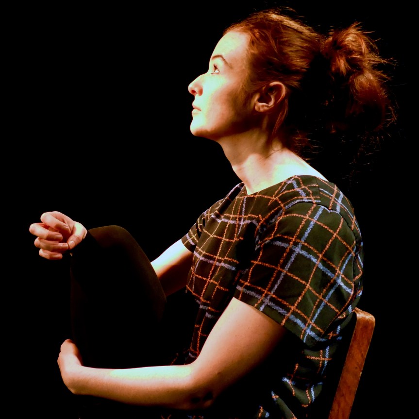 Photo of a young white woman swimming with her fingers and holding her knee whilst sat on a chair gazing off into the distance
