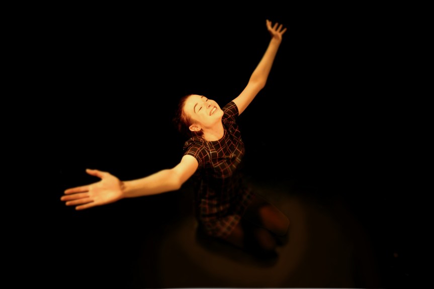 A young white woman with brown hair tied back, looking up and smiling, with her arms raised wide like wings